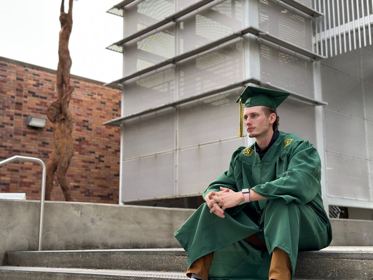 Graduate in a green cap and gown sitting on campus steps beside a bronze sculpture